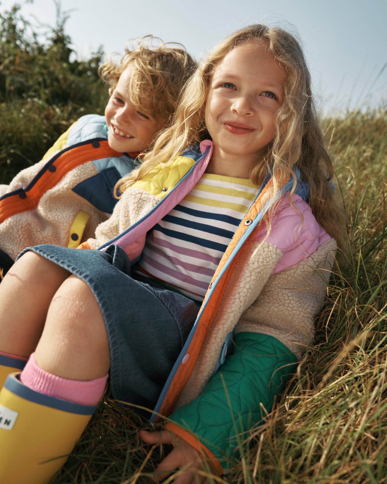 A close up  of a boy and a girl sat in a field wearing Boden coats.