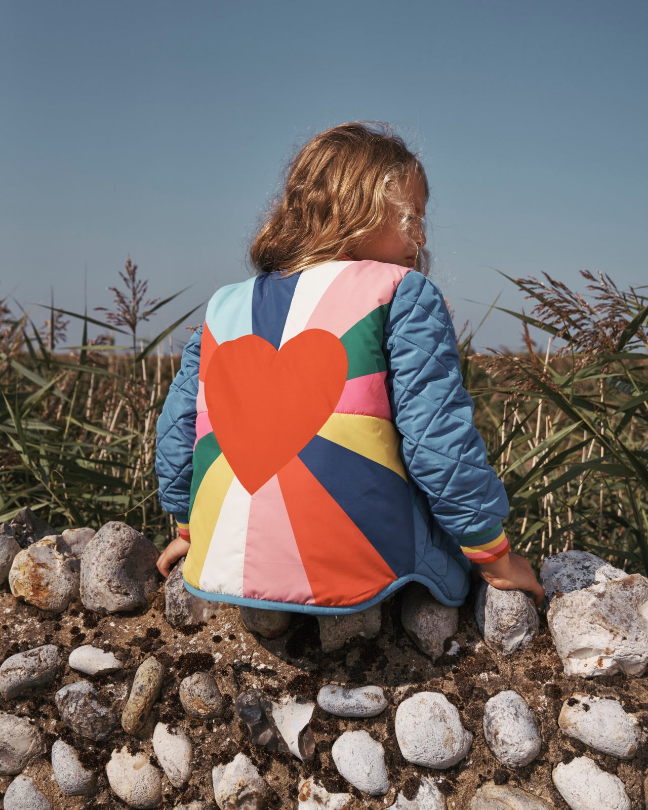 Girl sat with her back to the camera on a stone wall wearing a Boden jacket with a heart design