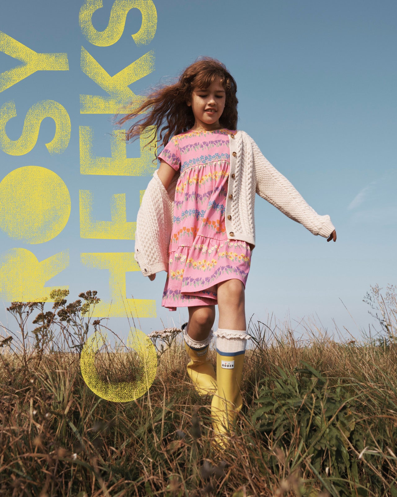 Girl running through a field wearing a Boden dress, cardigan and wellies.