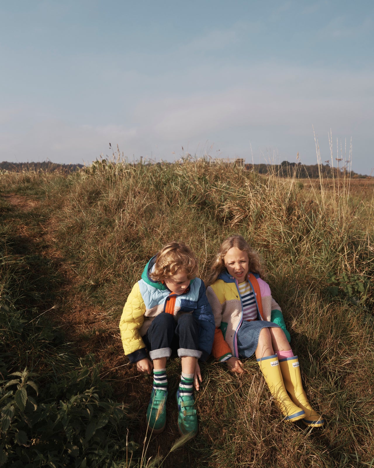 A boy and a girl sat in a field wearing Boden clothing.