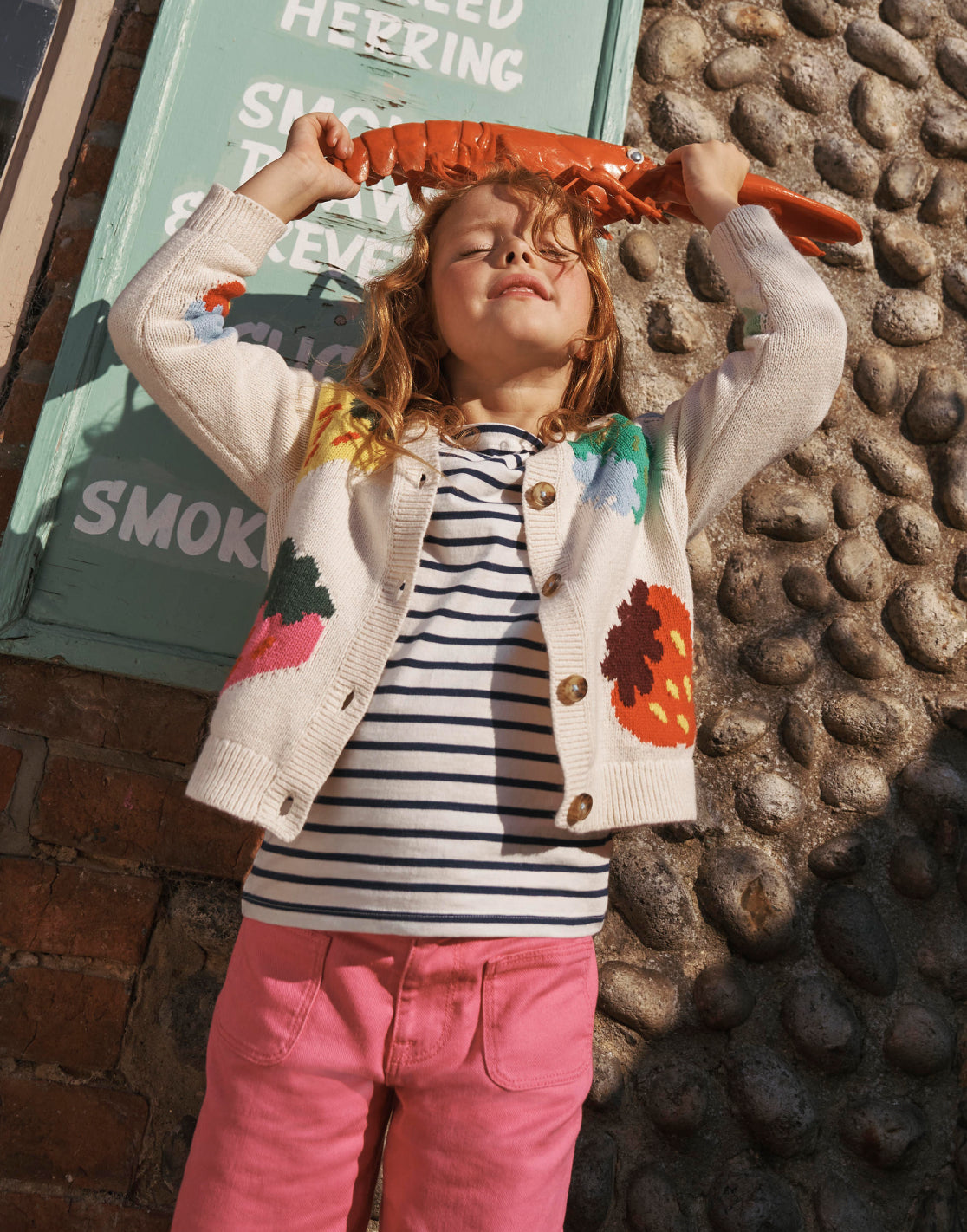 Girl holding a plastic lobster wearing a Boden knit cardigan with a colorful strawberry design.